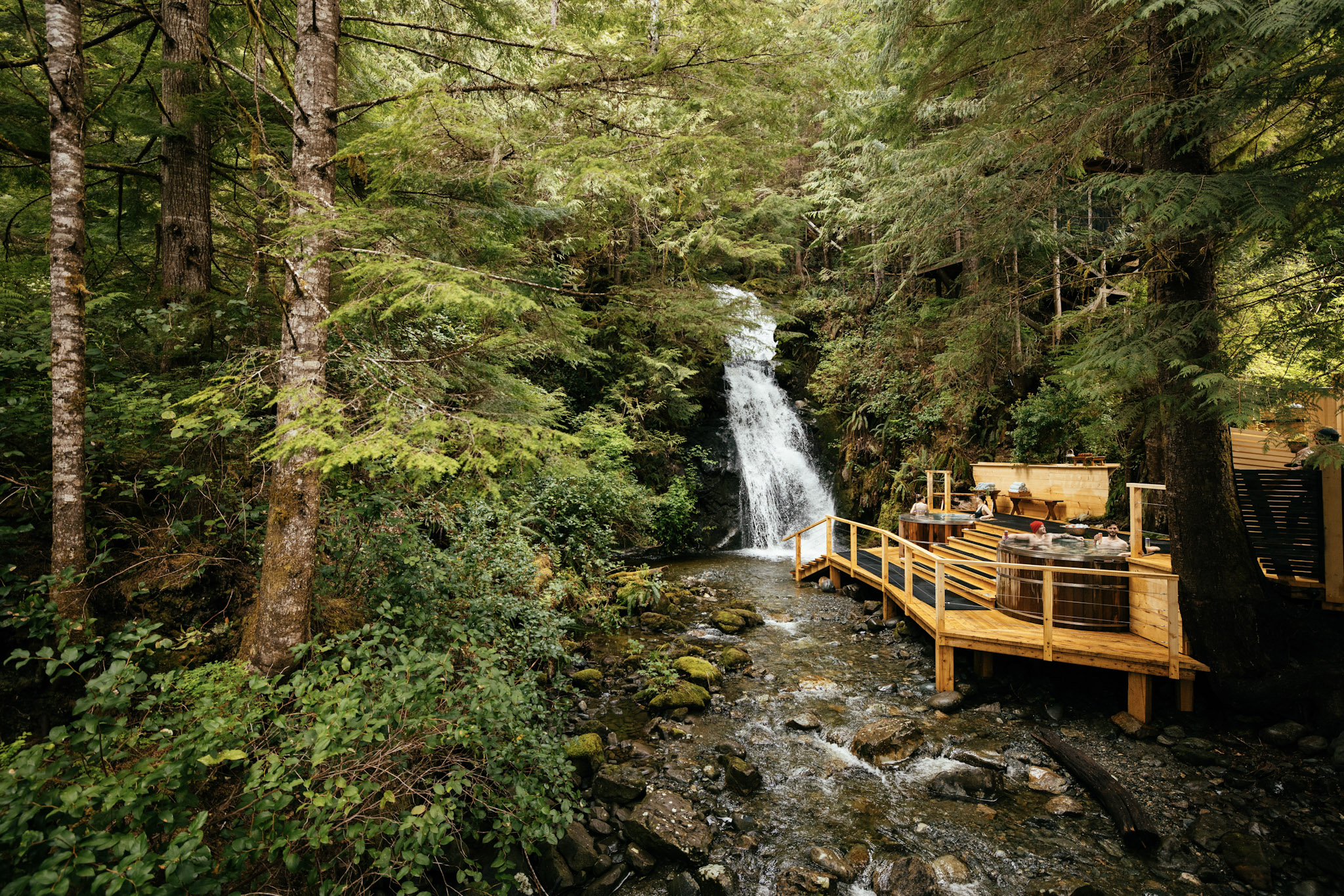 Cedar hot tubs on a wooden deck at the base of Nimmo Bay's waterfall, surrounded by rainforest