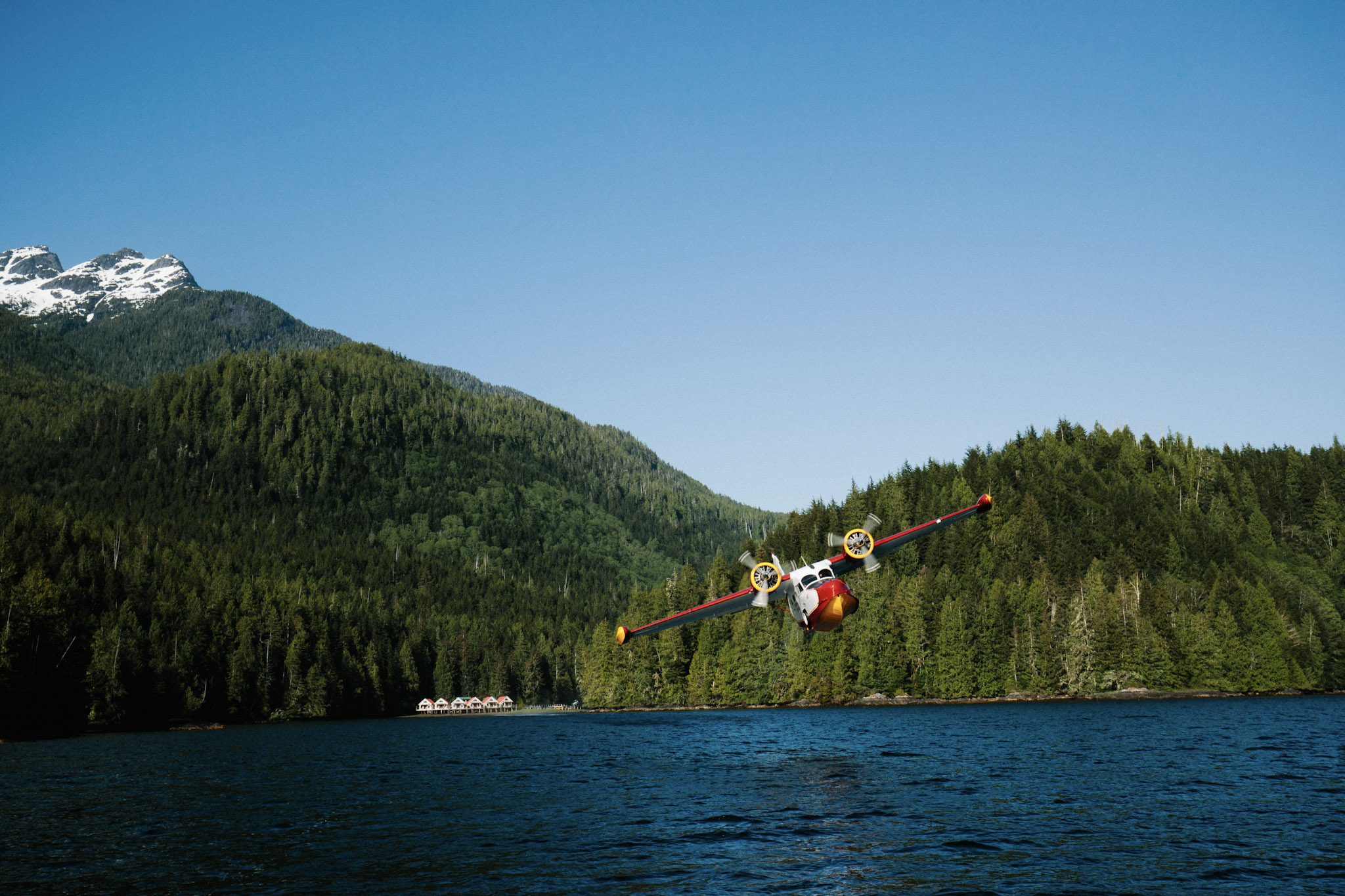 A floatplane banks over the inlet at Nimmo Bay, the resort's shoreline cabins below and snow-capped peaks beyond