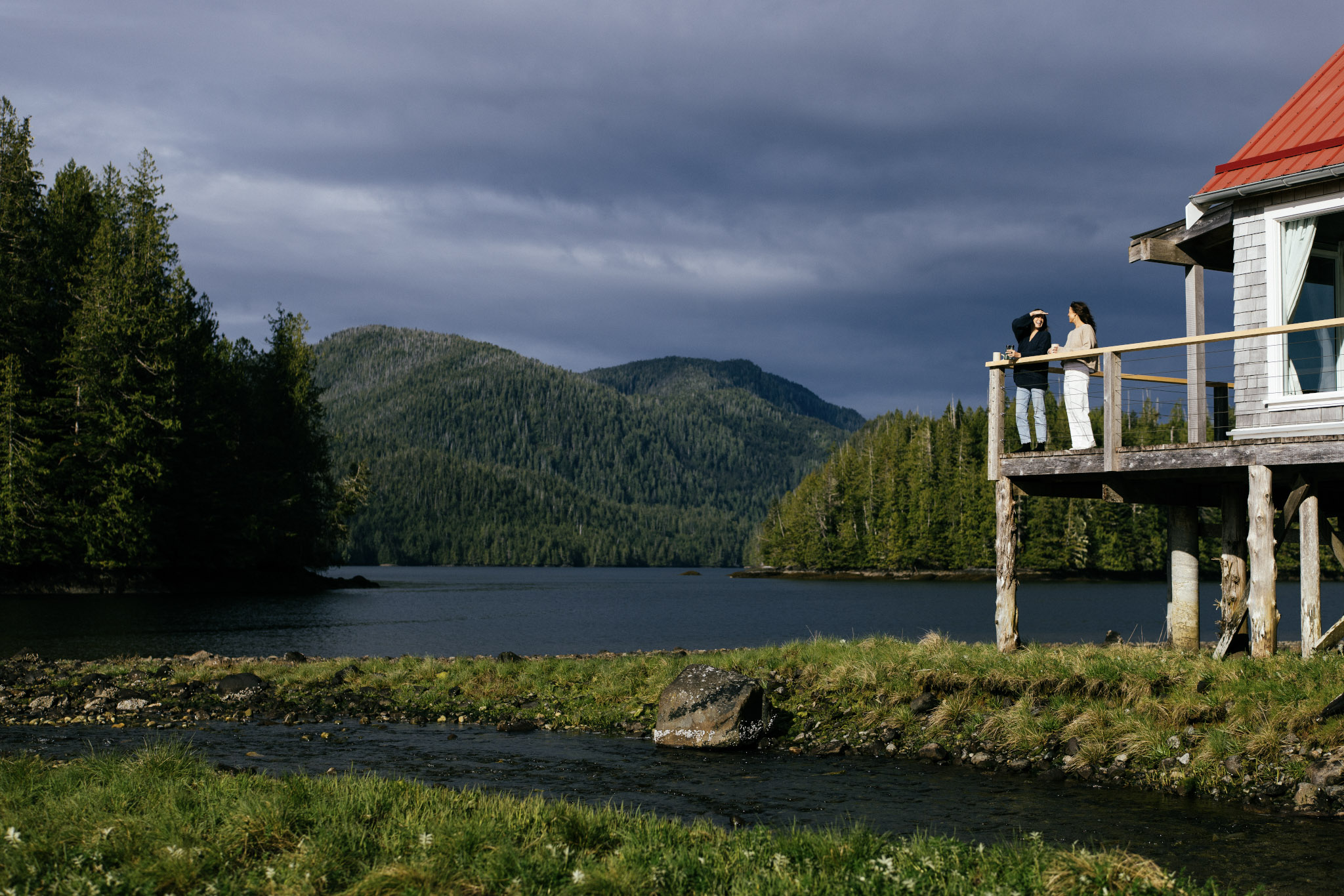 Two guests on the balcony of a stilted cabin at Nimmo Bay, looking out over the inlet under storm light