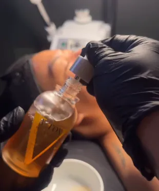 Hands wearing black gloves holding and pouring amber-colored liquid from a dropper bottle near a person's face blurred in the background.