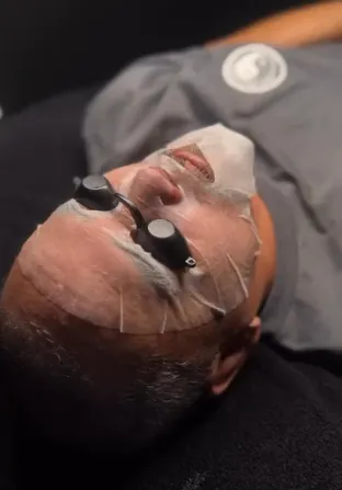 Person lying down with a transparent facial mask and black protective goggles during a skincare treatment.