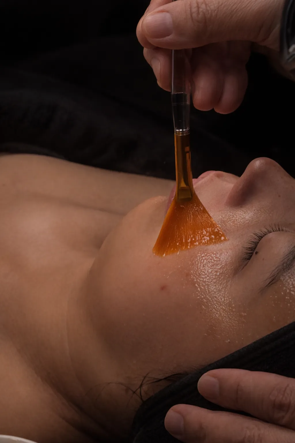 Close-up of a person receiving a facial treatment with a brush applying an orange gel mask.