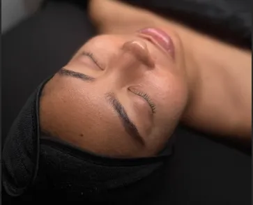 Close-up of a woman with closed eyes and smooth skin, wearing a black head wrap, lying down on a black surface.