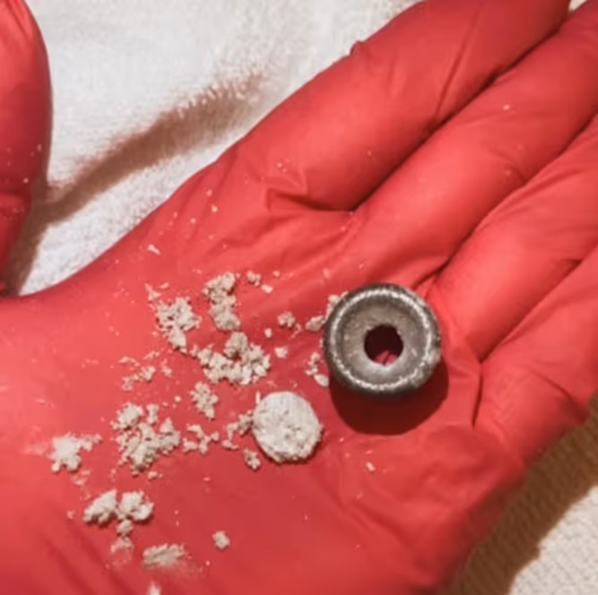 Close-up of a red-gloved hand holding a metallic cylindrical object with a hole and white powdery substance scattered next to it.