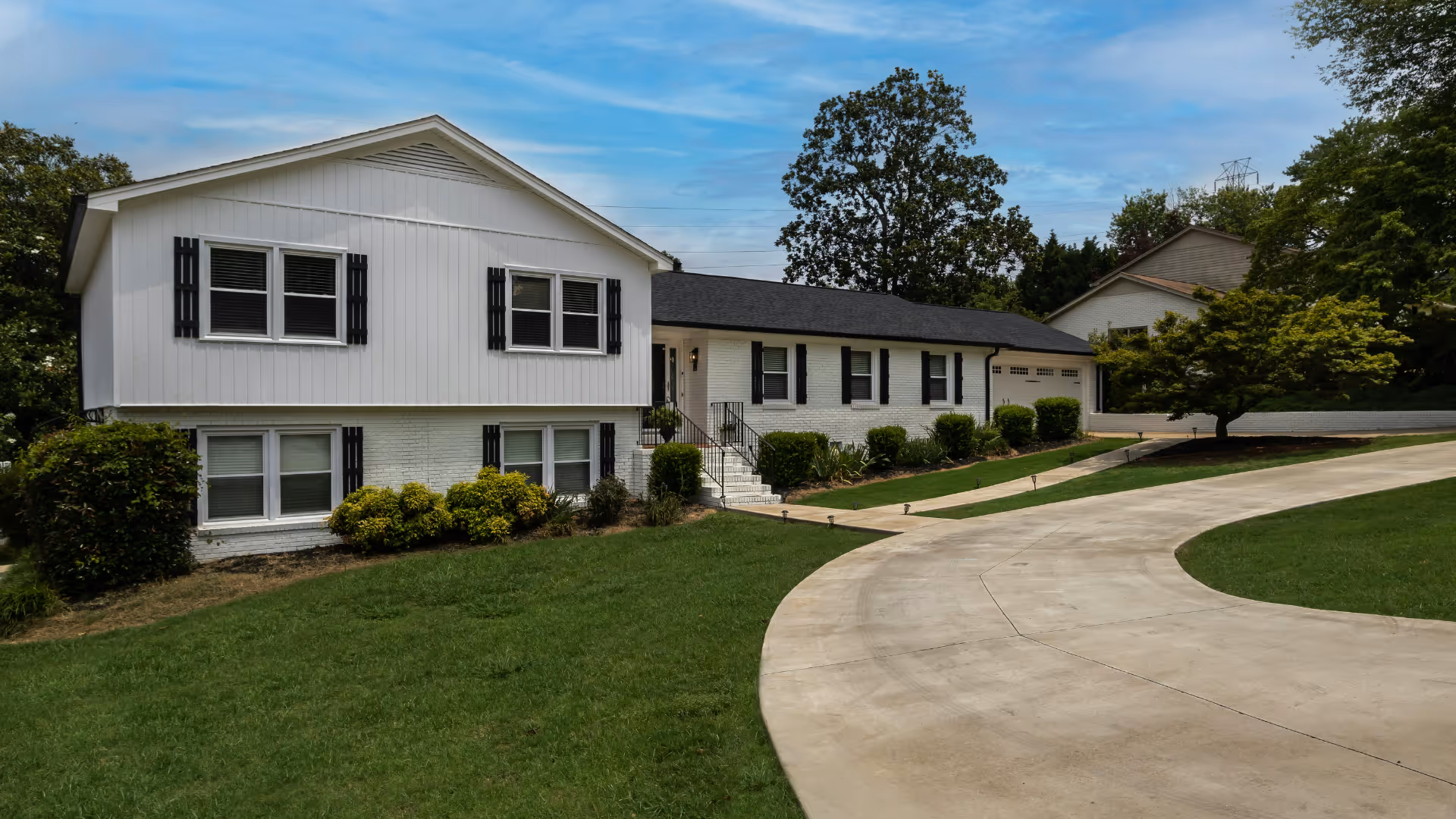 White multi-level house with black shutters, curved concrete driveway, and green lawn under a blue sky.