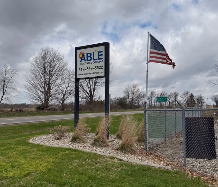 Outdoor sign for Able Heating & Cooling with phone number and website, near a US flag and road.