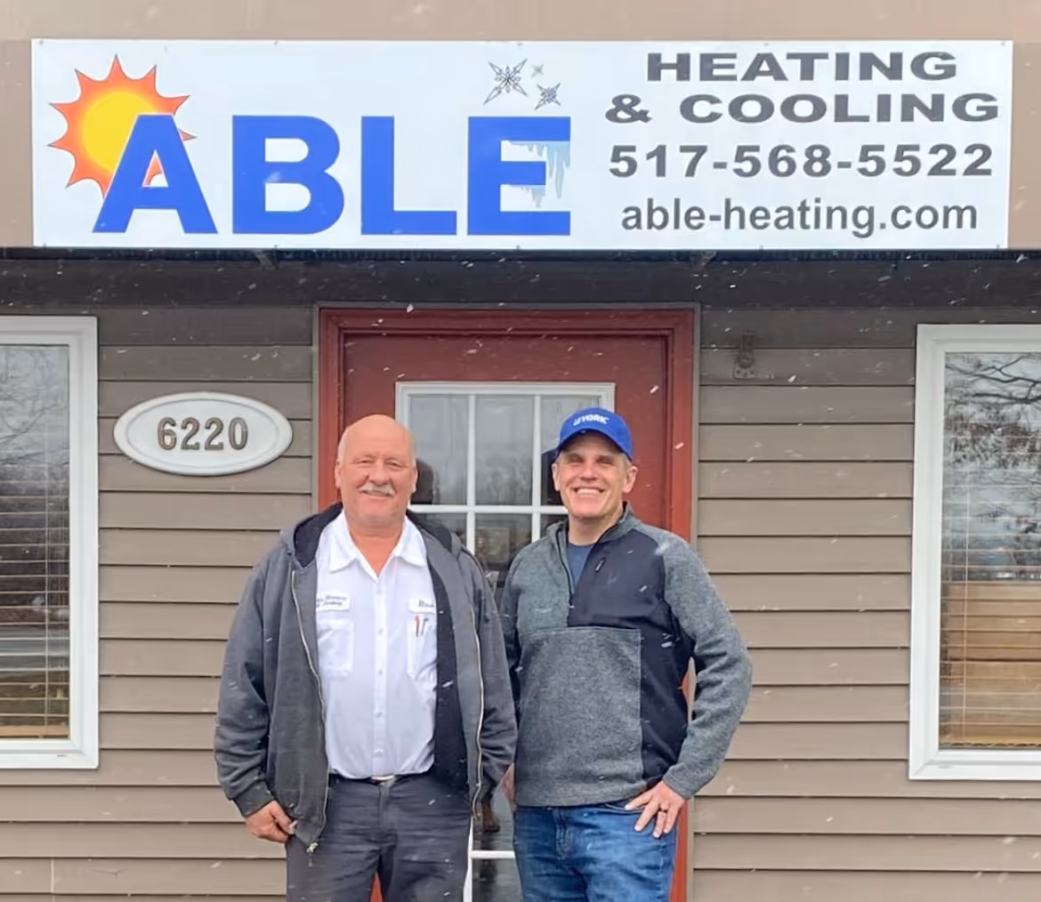 Two men standing outside a building under a sign for Able Heating & Cooling with phone number and website.