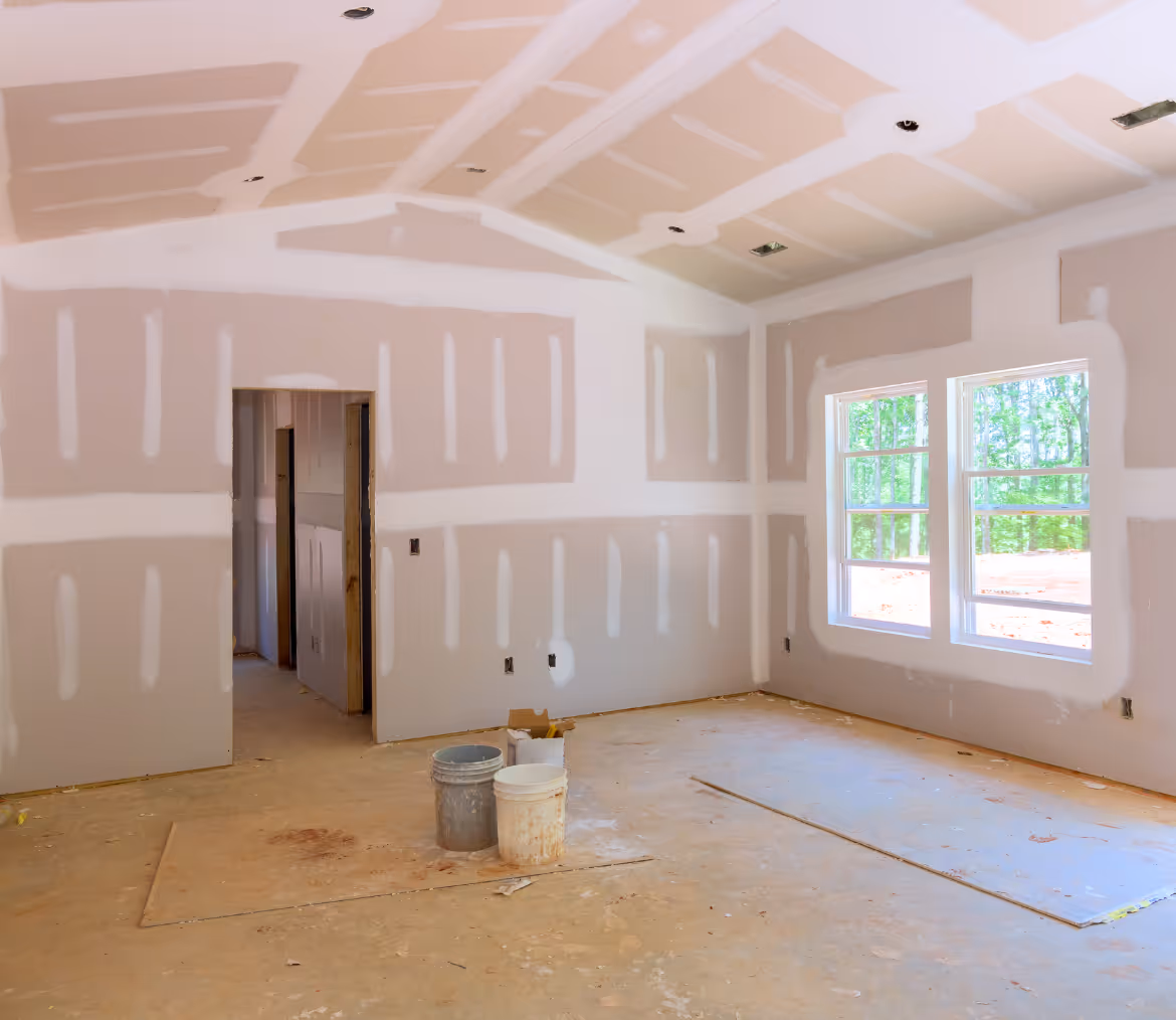 Interior of an unfinished room with drywall installed, two windows, and construction buckets on the floor.