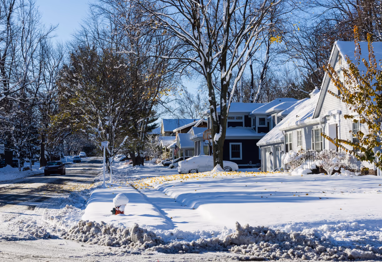 Residential street with houses and trees covered in fresh snow under a clear blue sky.