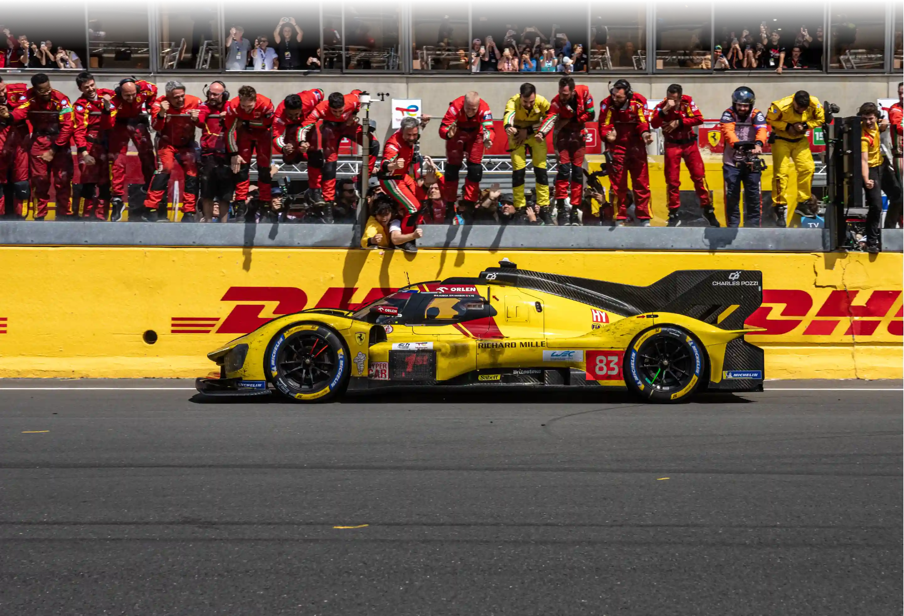 Yellow and black race car number 83 passes the pit wall while team members in red and yellow uniforms cheer from above.