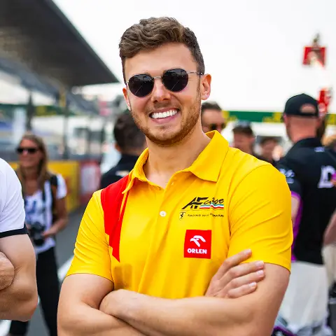 Smiling man wearing sunglasses and a yellow racing team polo shirt with ORLEN logo at a race event.