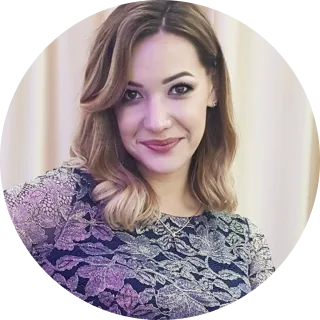 Smiling woman with shoulder-length wavy hair wearing a lace floral patterned top against a neutral background.