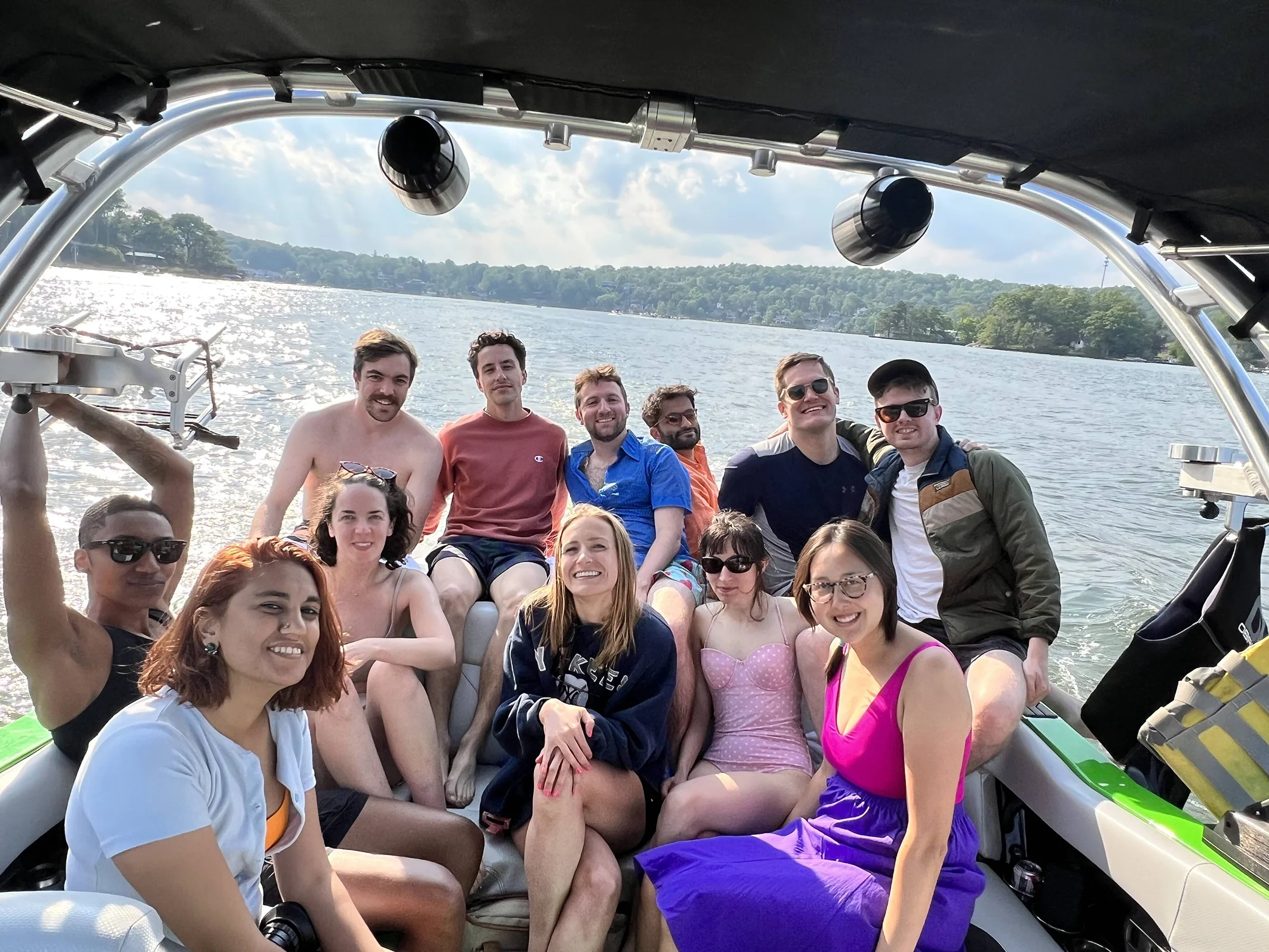 Group of eleven young adults smiling and sitting closely together on a boat on a sunny lake, with greenery and houses visible in the background.