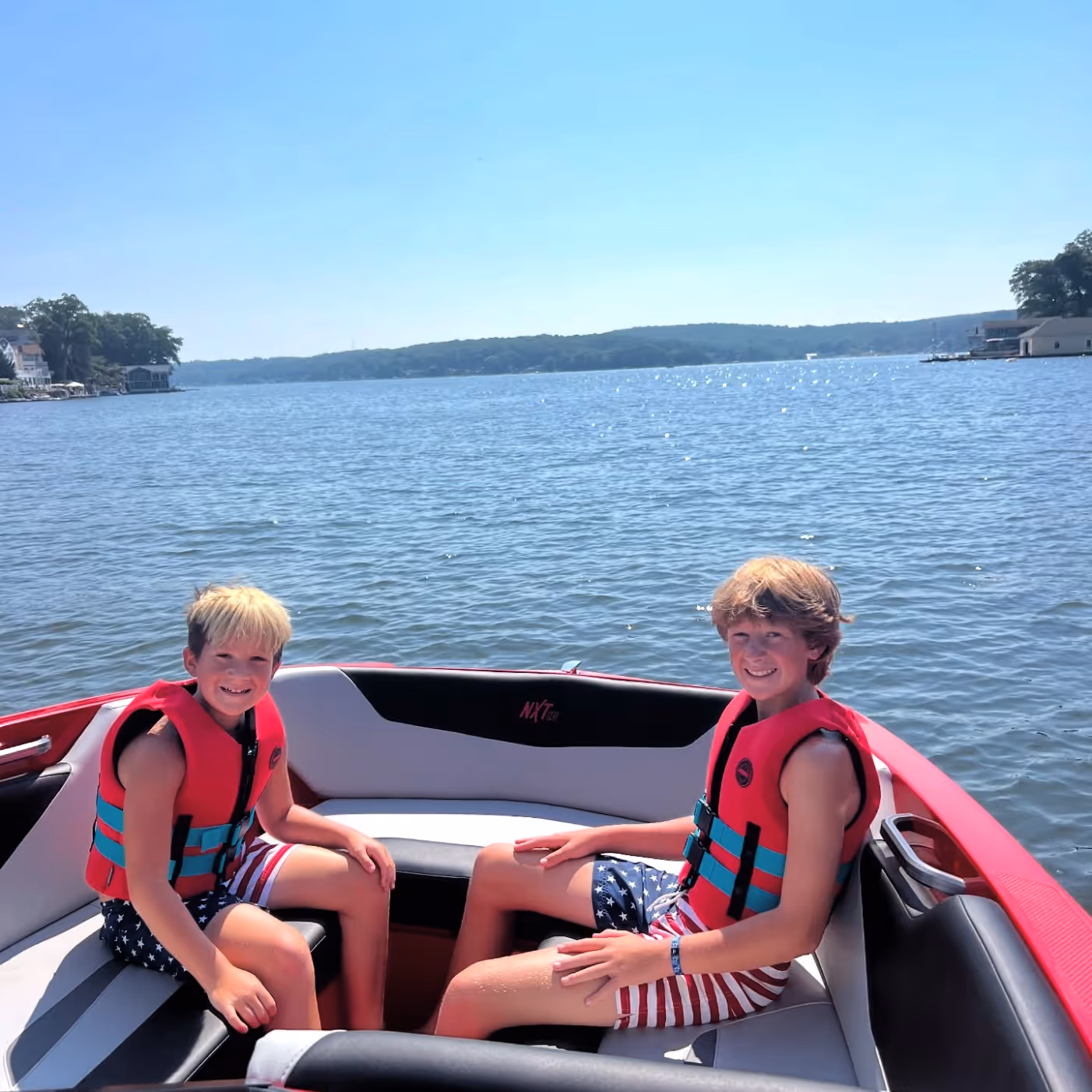 Two boys wearing red life jackets and American flag-themed swim trunks sitting and smiling on a boat on a sunny lake.