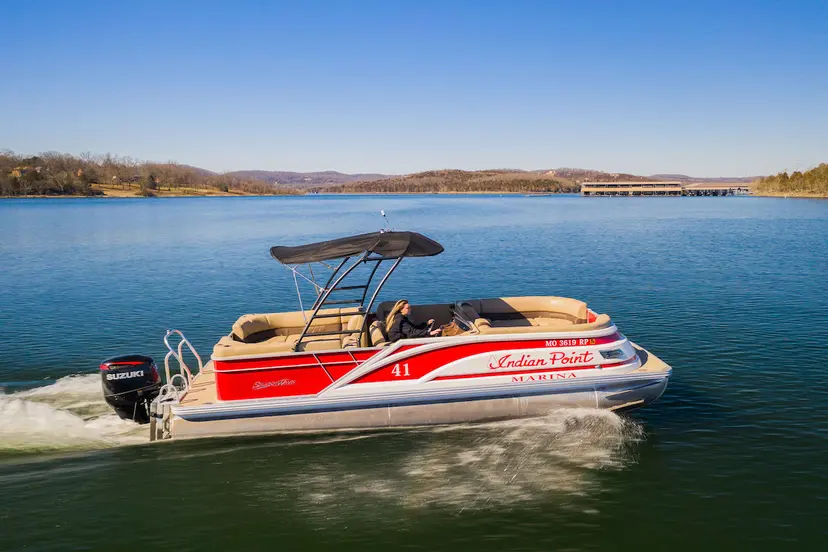 Five people relaxing on a South Bay pontoon boat with red and black accents on a calm lake near houses and trees under a clear blue sky.
