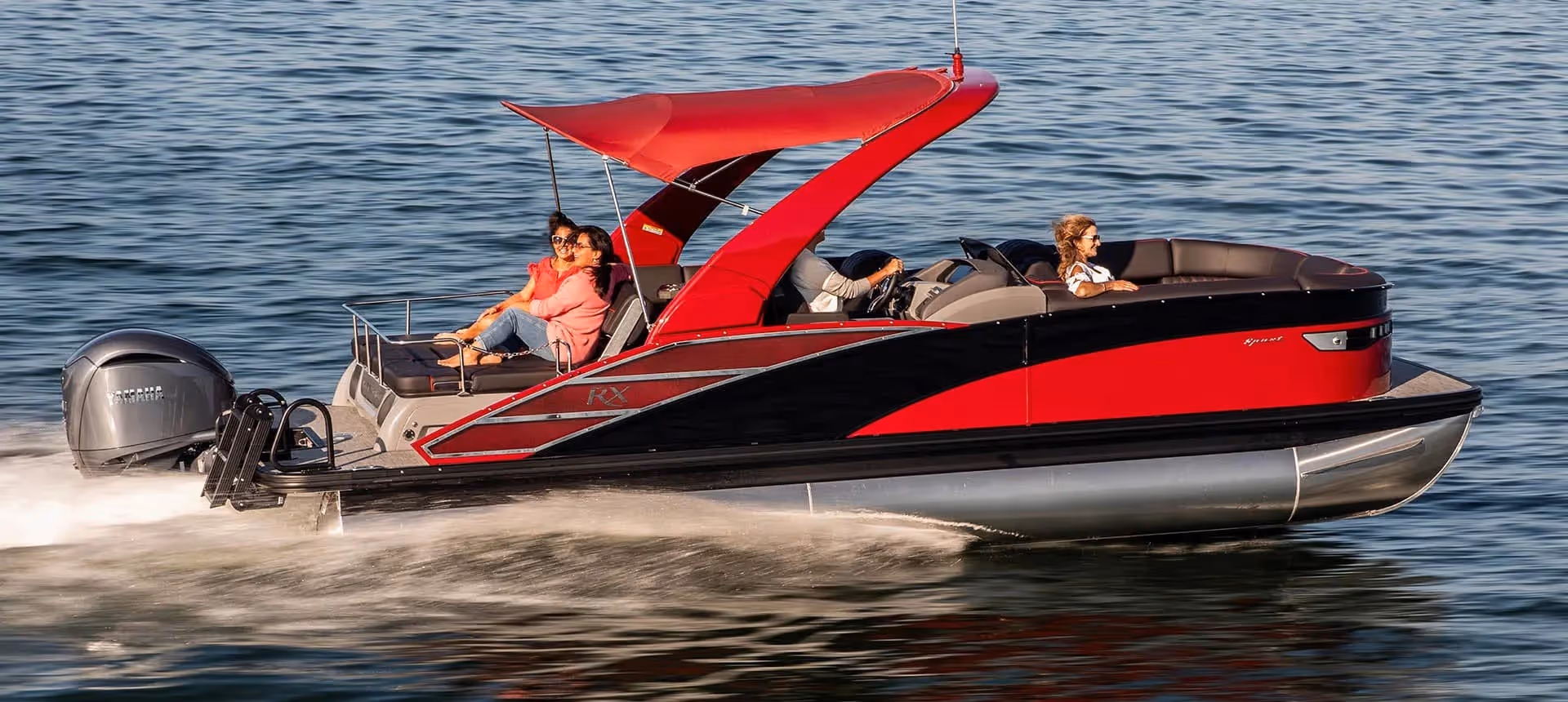 Five people relaxing on a South Bay pontoon boat with red and black accents on a calm lake near houses and trees under a clear blue sky.