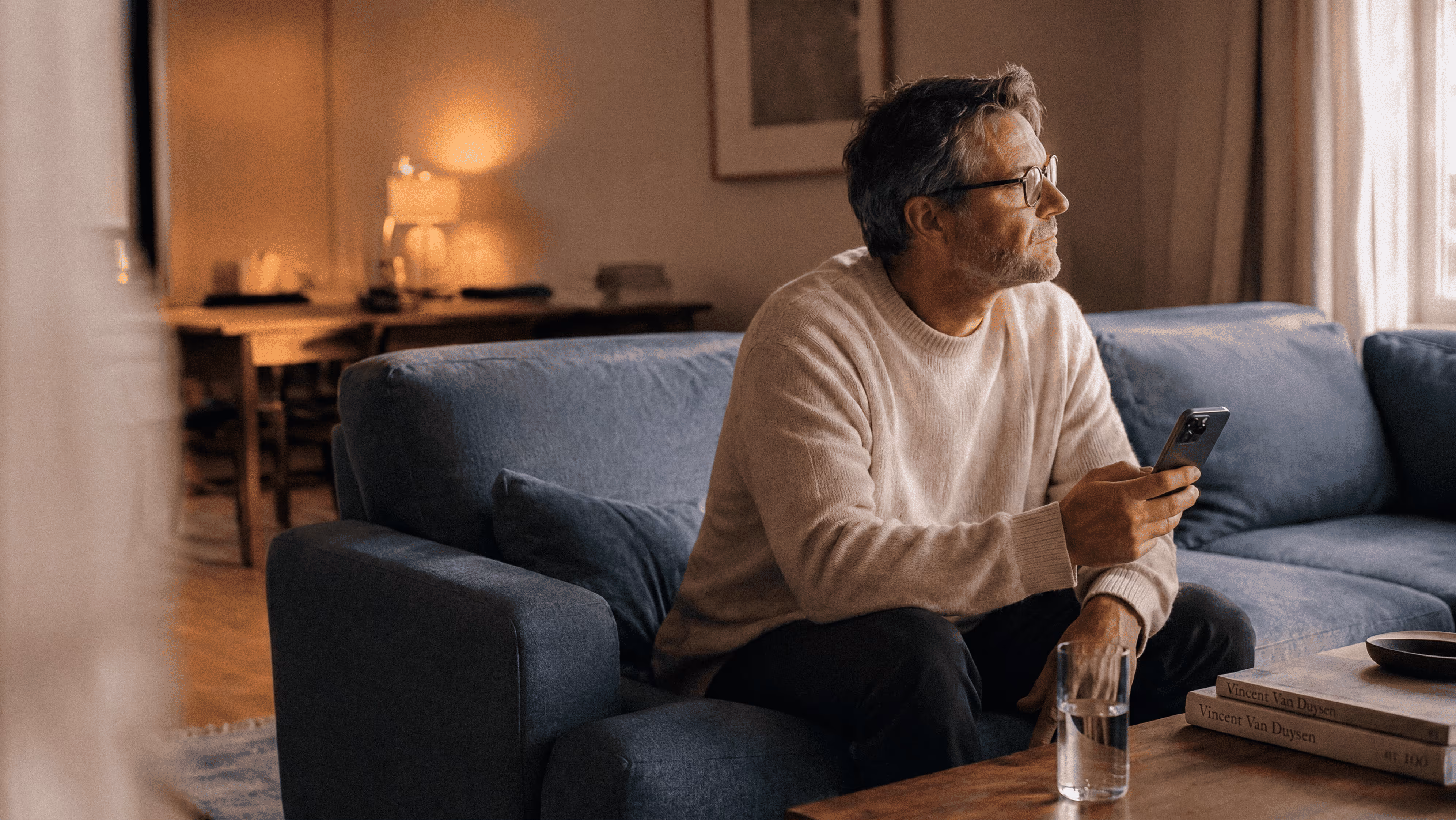 Älterer Mann mit Brille sitzt auf grauem Sofa und hält Smartphone in warmer Inneneinrichtung.