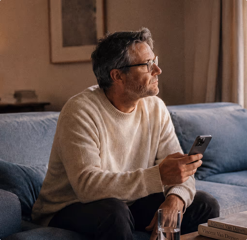 Älterer Mann mit Brille sitzt auf Couch und hält Smartphone in der Hand.