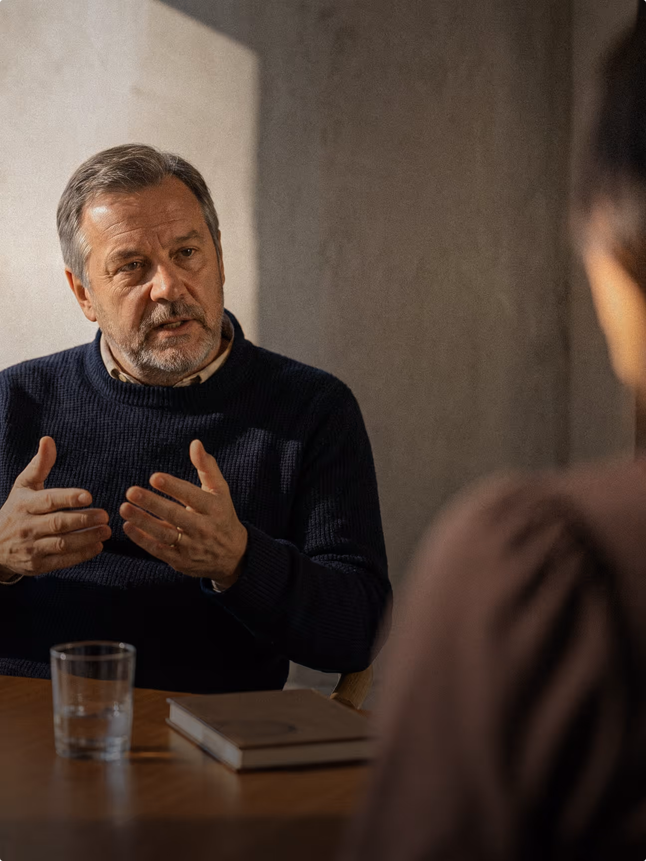 Älterer Mann im dunklen Pullover spricht gestikulierend mit einer Person am Tisch.