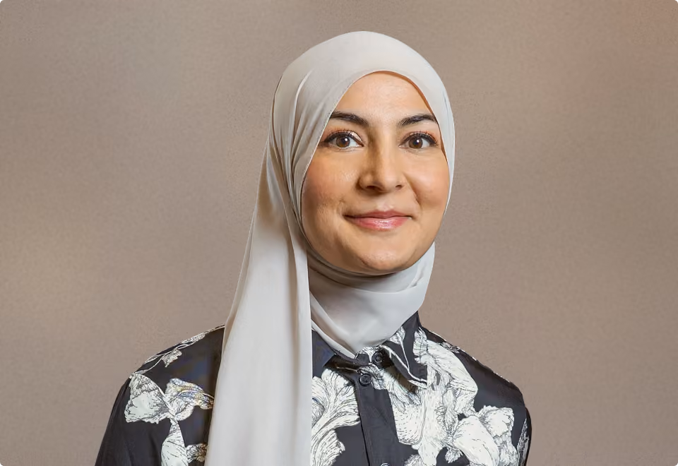 Porträt einer Frau mit weißem Hijab und gemustertem schwarzen Hemd, lächelnd.
