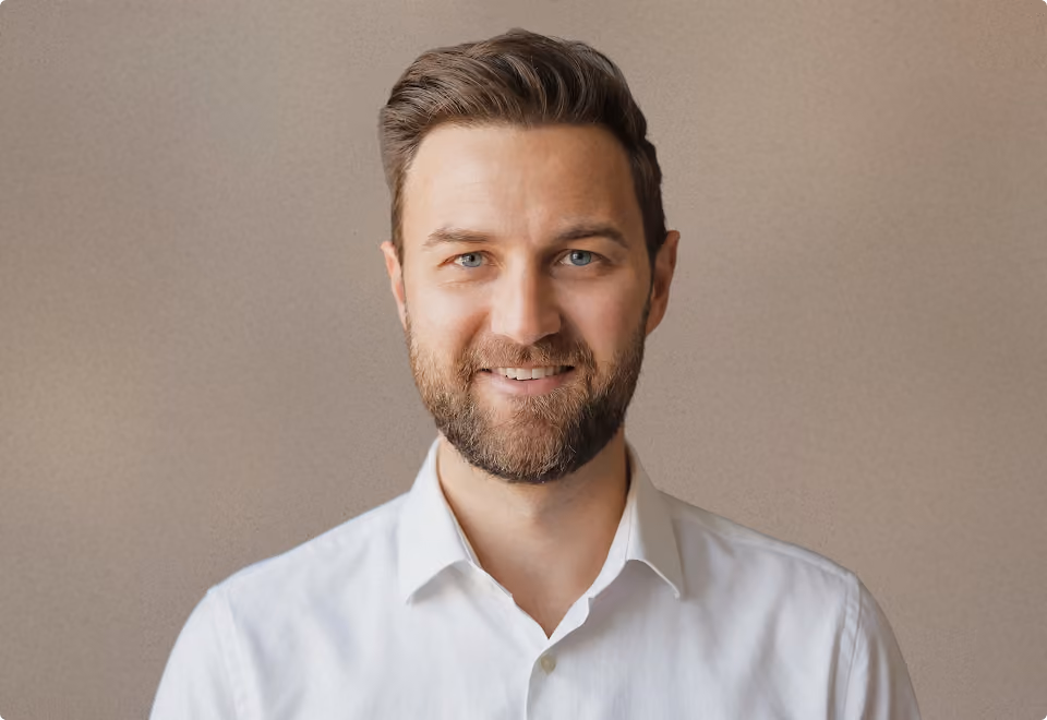Portrait eines lächelnden Mannes mit Bart in weißem Hemd vor neutralem Hintergrund.
