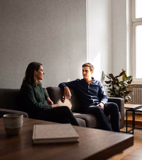 Zwei Personen sitzen sich auf einem grauen Sofa in einem hellen Wohnzimmer gegenüber.