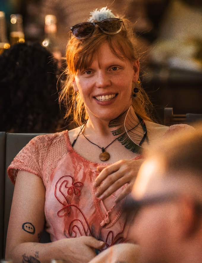 Frau mit roten Haaren, Sonnenbrille und rosa Shirt lächelt in warmer Atmosphäre.