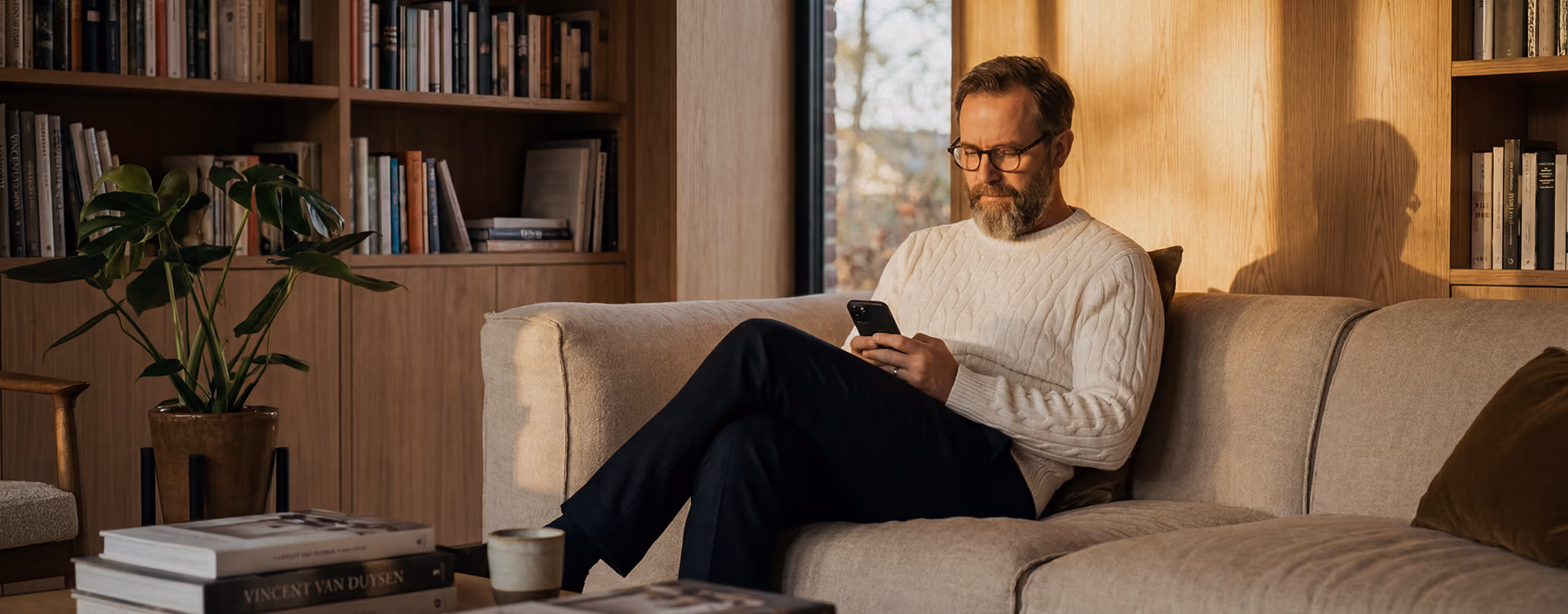 Mann sitzt auf Sofa und schaut auf sein Smartphone in gemütlichem Wohnzimmer mit Bücherregalen.