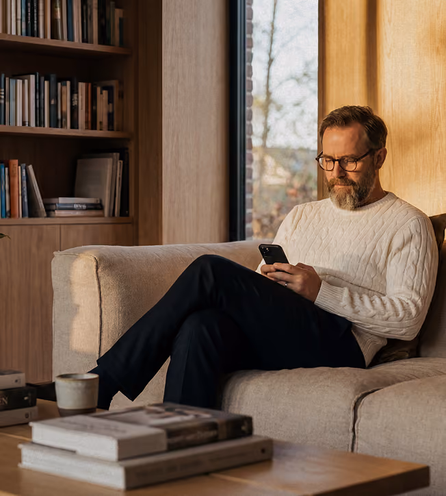 Mann mit Brille sitzt auf Sofa, schaut auf Smartphone, Bücherregal im Hintergrund