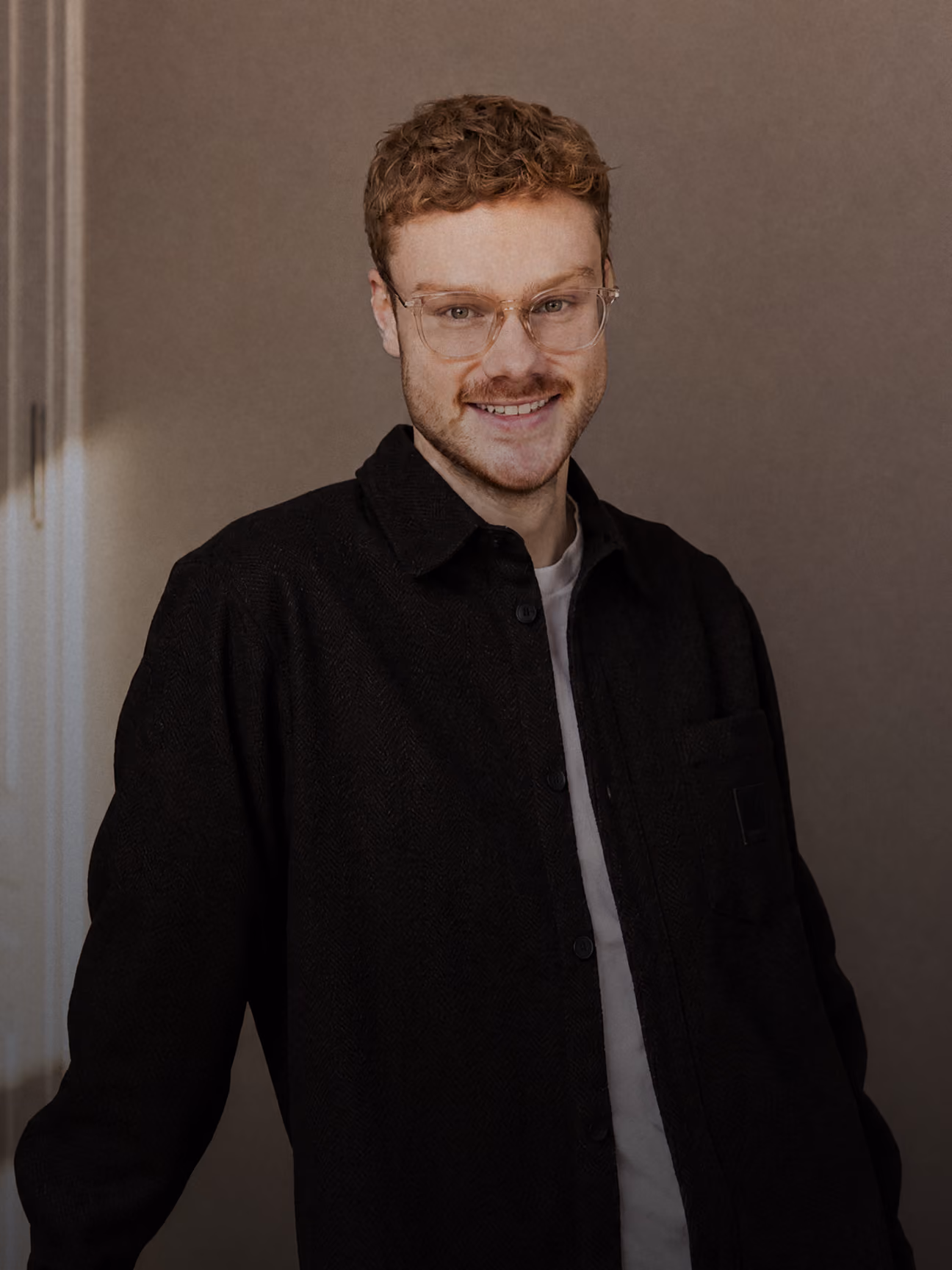 Junger Mann mit Brille, braunem lockigem Haar und schwarzer Jacke lächelt