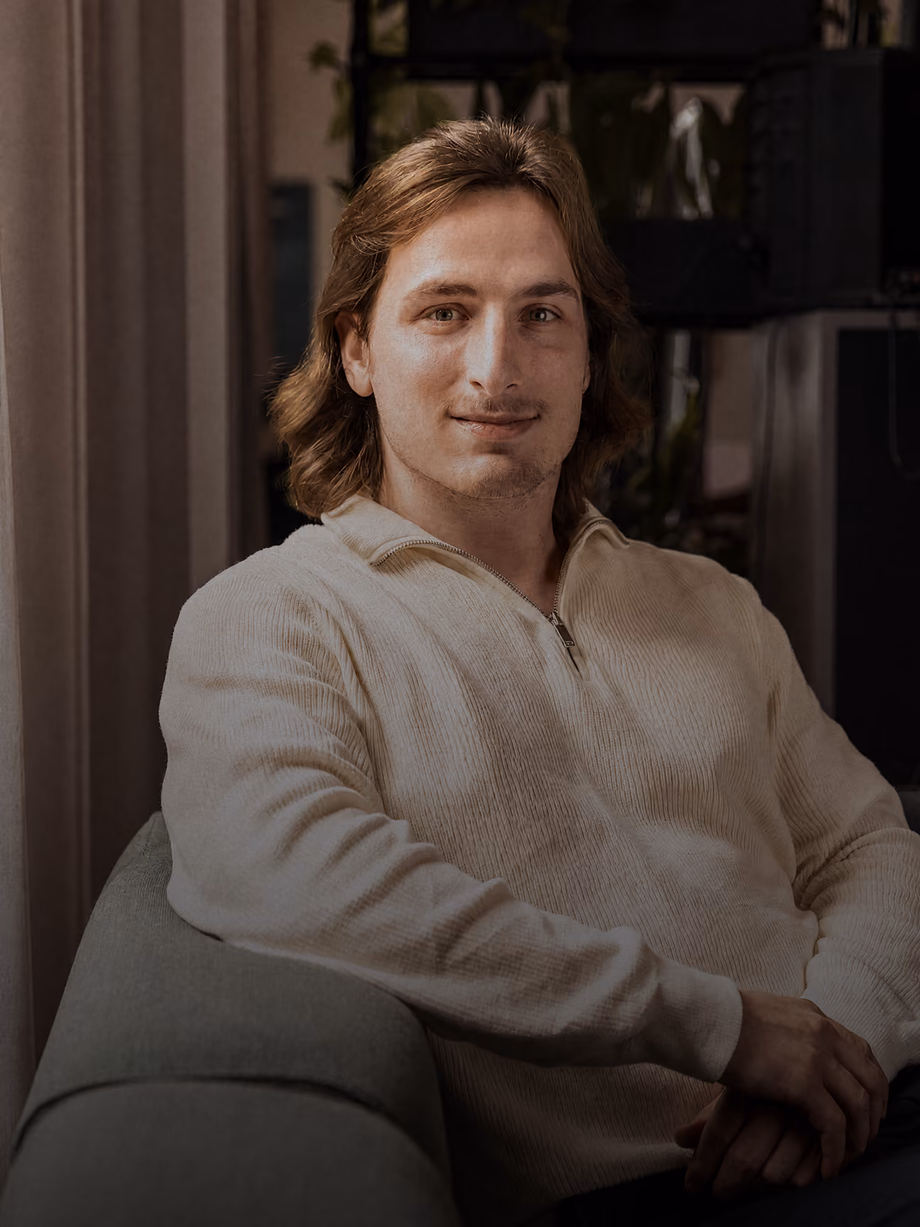 Mann mit schulterlangem braunem Haar sitzt in cremefarbenem Pullover.