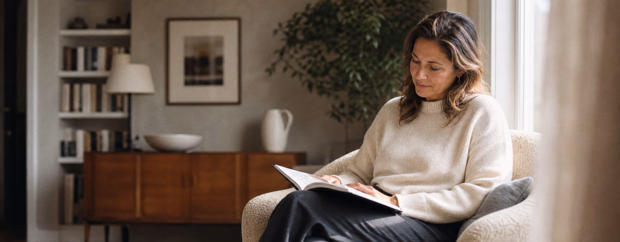Frau in hellem Pullover sitzt im modernen Wohnzimmer und liest konzentriert eine Zeitschrift.