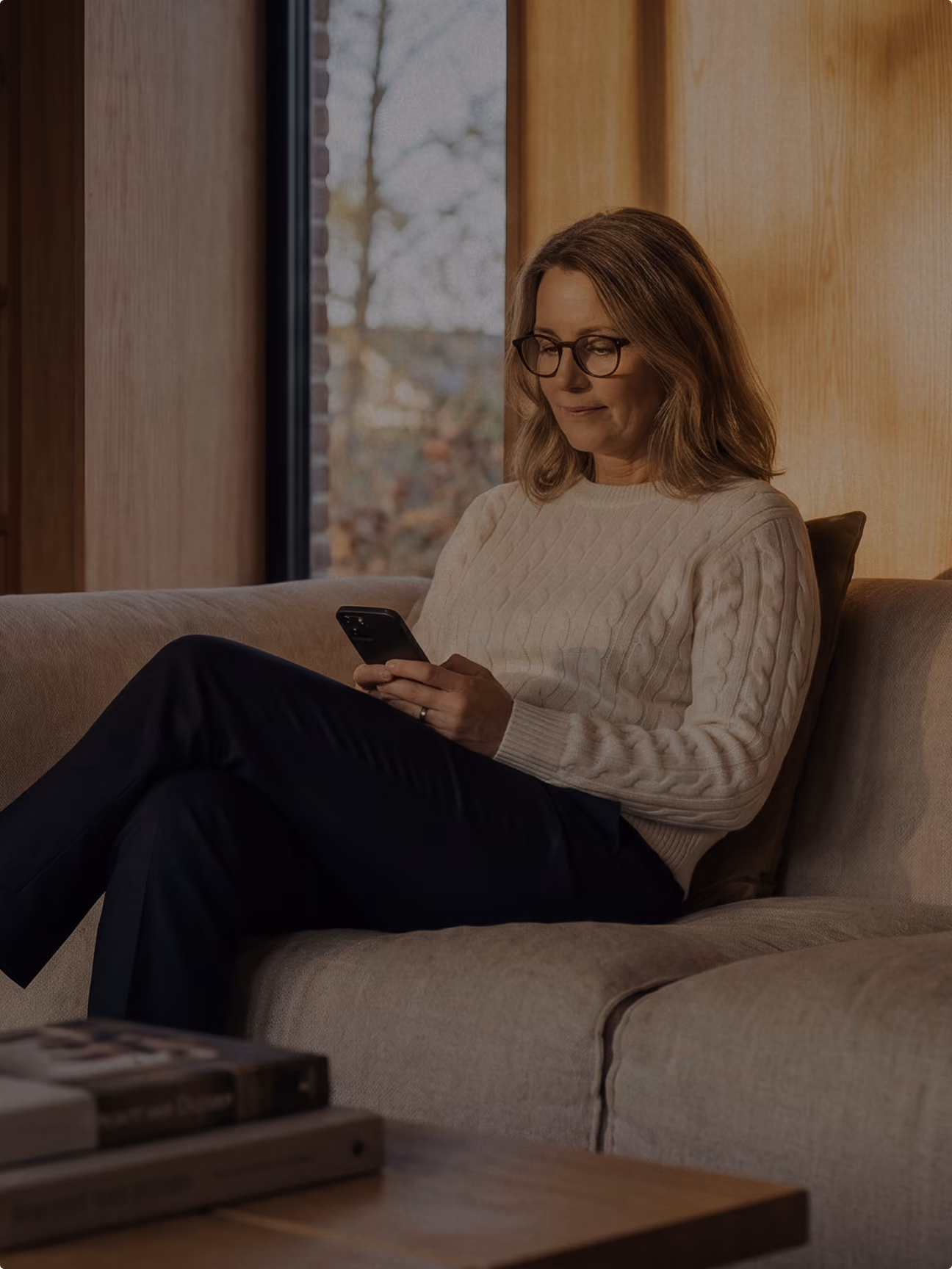 Frau mit Brille sitzt auf Couch und schaut auf ihr Smartphone in warmer Inneneinrichtung.