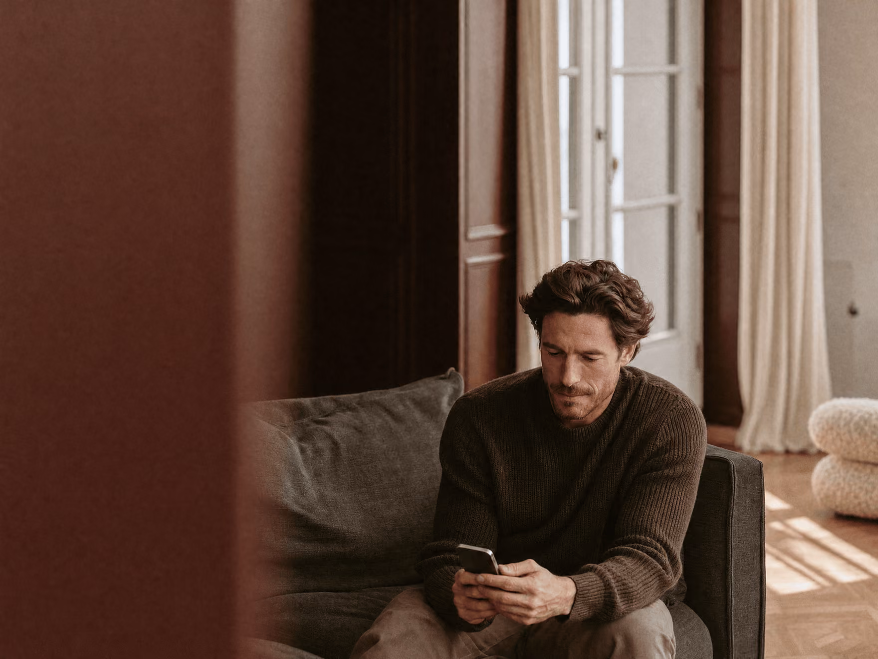 Mann sitzt auf Sofa und schaut auf sein Smartphone in hellem, modernem Wohnzimmer.