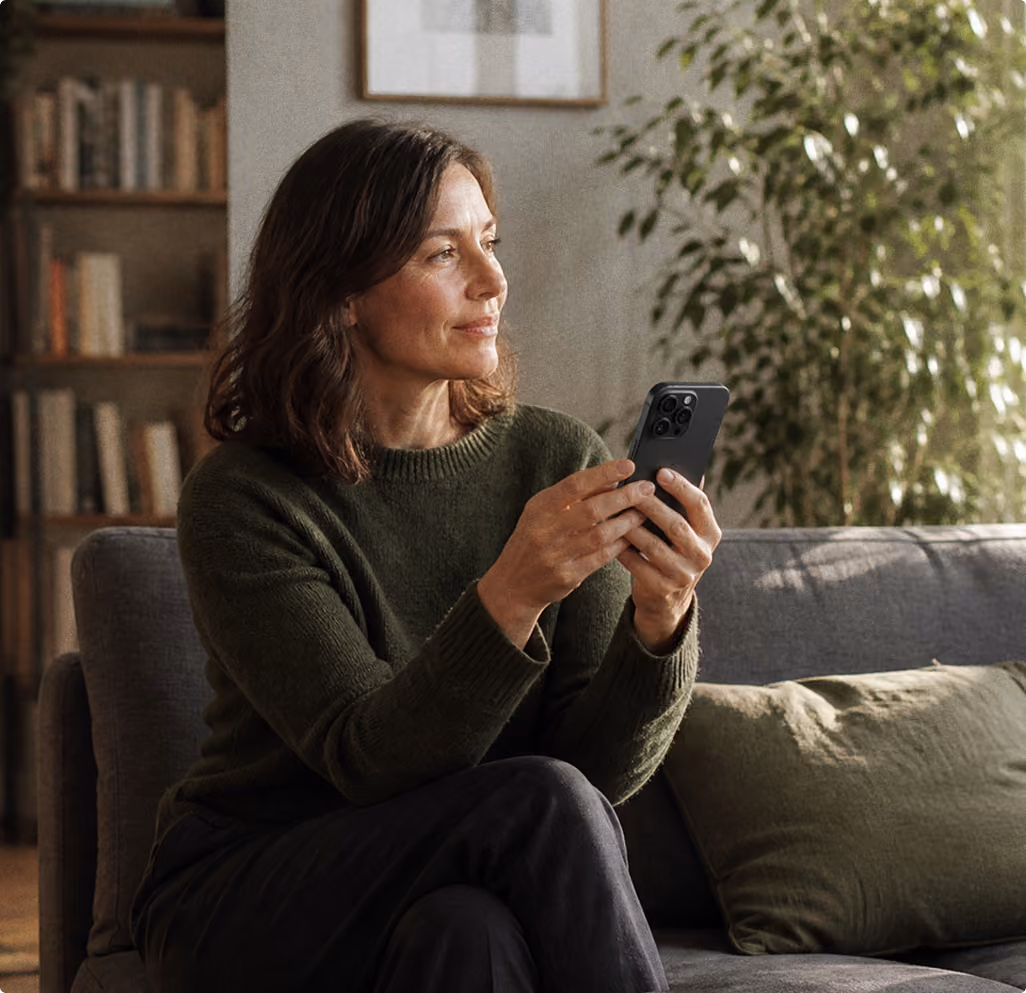 Frau sitzt auf Sofa und hält Smartphone in grünem Pullover in gemütlichem Wohnzimmer.