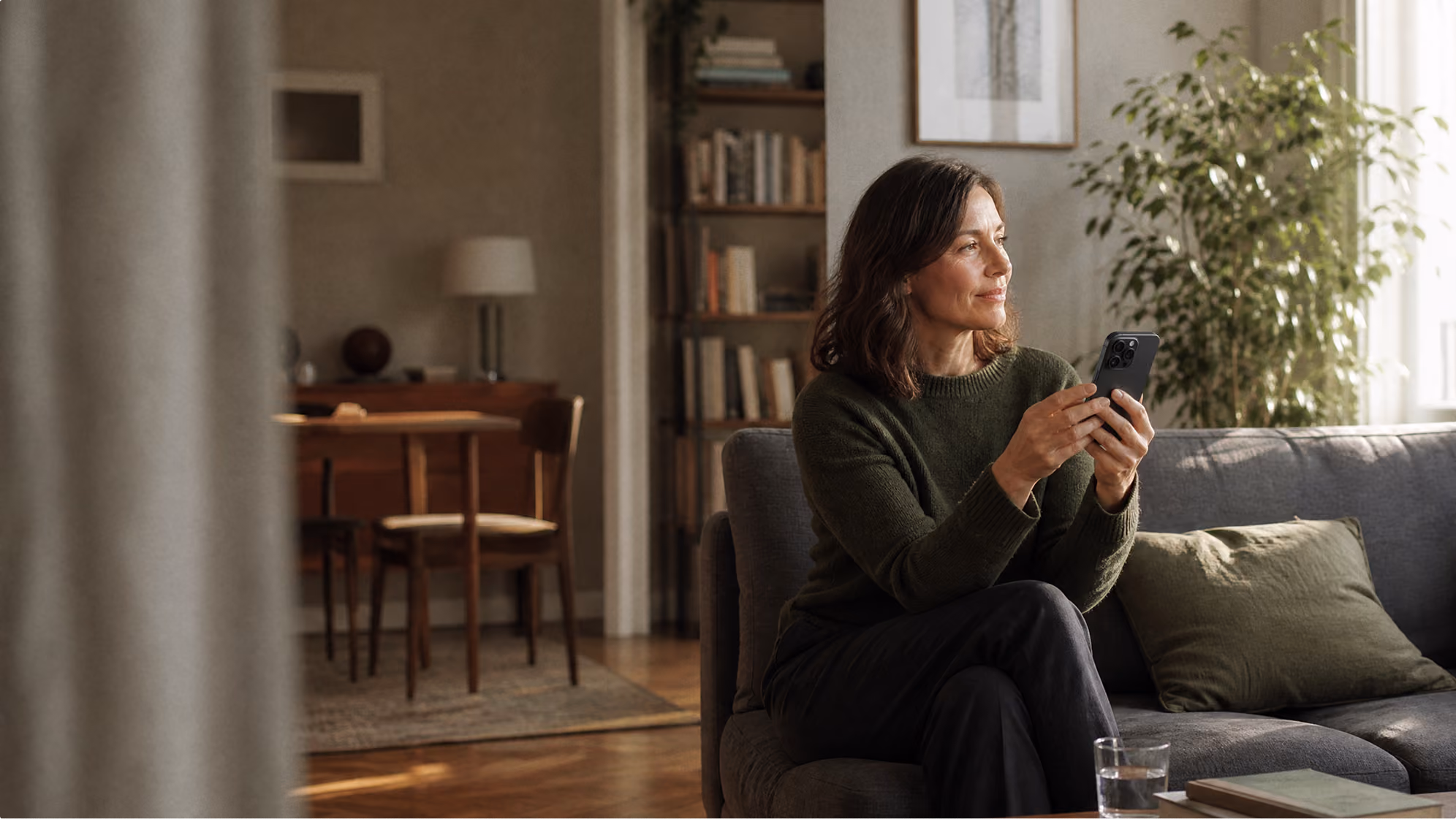 Frau sitzt auf Sofa und hält Smartphone in moderner, heller Wohnzimmerumgebung mit Pflanzen.