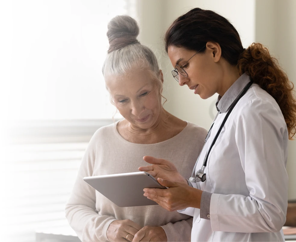 Female doctor showing information on a tablet to an elderly woman in a bright room.