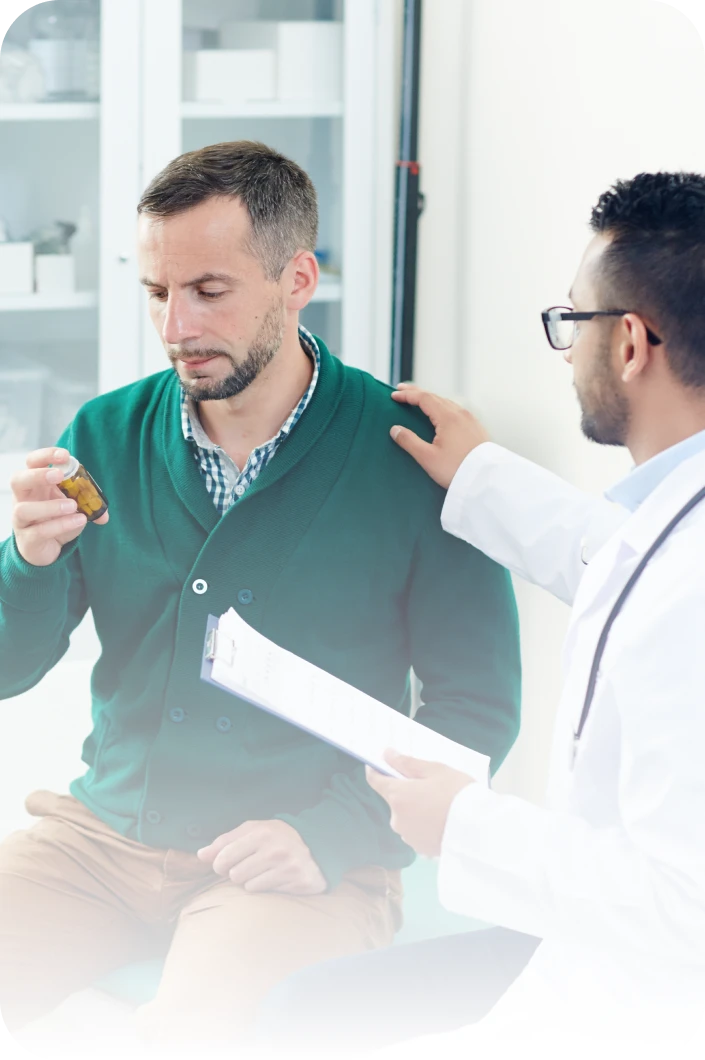 A doctor placing a reassuring hand on a man's shoulder while the man looks at a prescription medicine bottle.