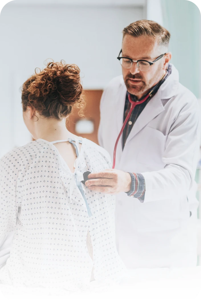 Male doctor in white coat using a stethoscope to listen to a female patient's back in a hospital room.