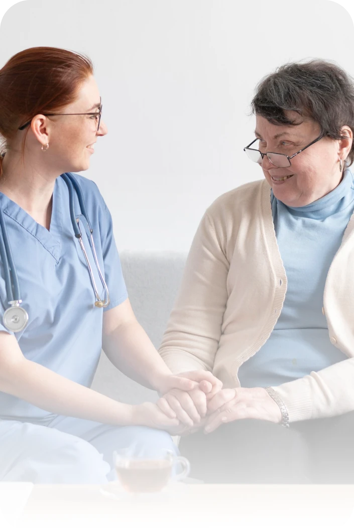 Nurse in blue scrubs holding hands and smiling at an older woman wearing glasses and a beige cardigan.