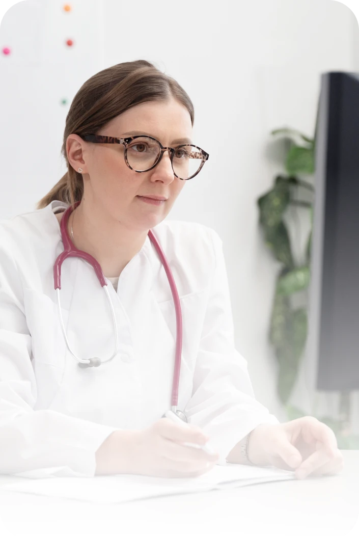 Female doctor wearing glasses and a white coat with a stethoscope around her neck, working at a desk.