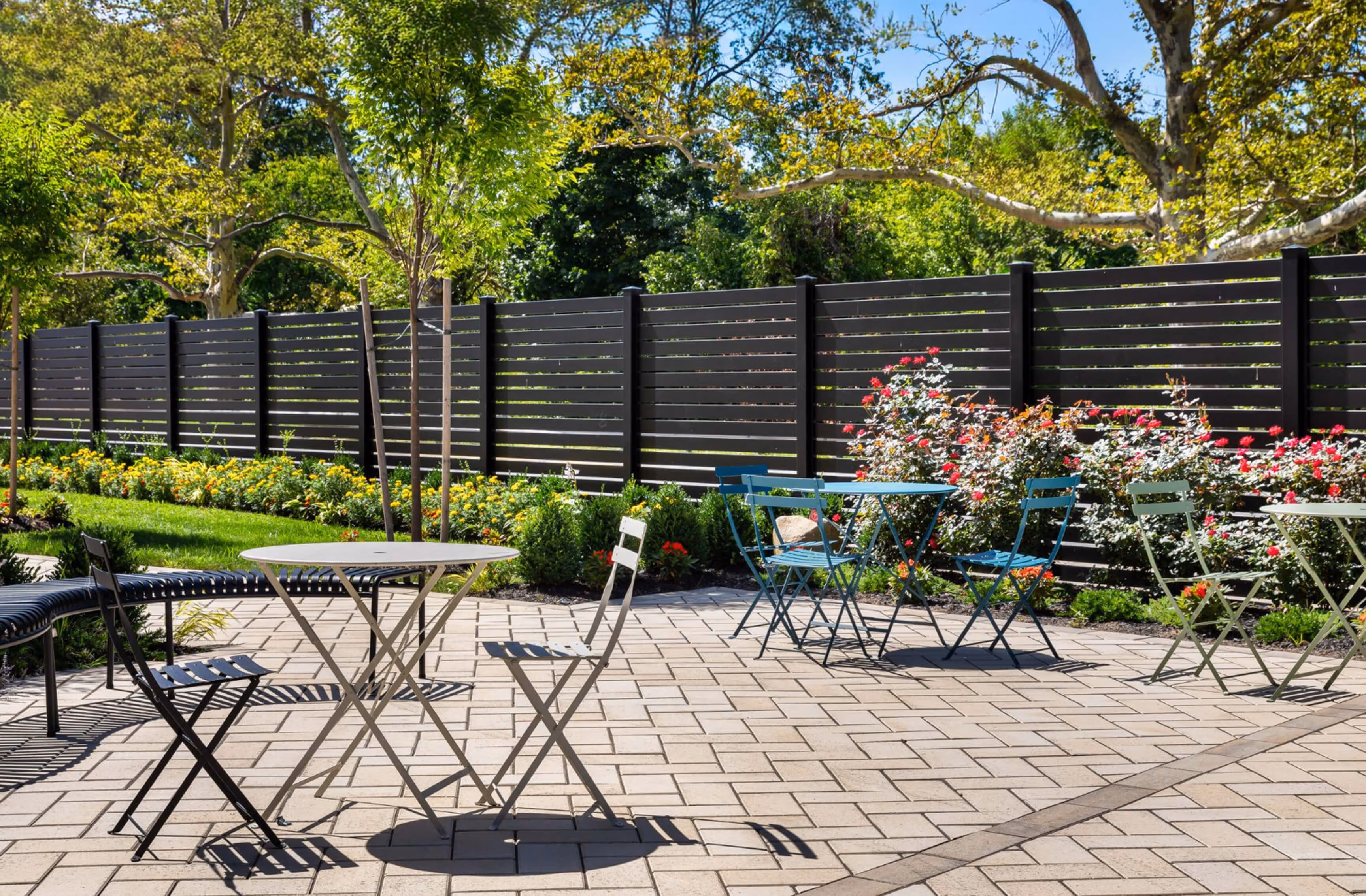 Outdoor patio with multiple small metal tables and chairs on a tiled floor next to a garden with flowers and a wooden fence.