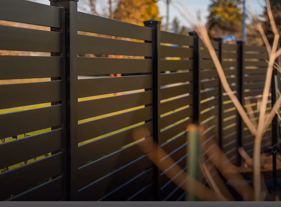 Dark brown horizontal slat privacy fence with spaced gaps between slats, outdoors with blurred trees in the background.
