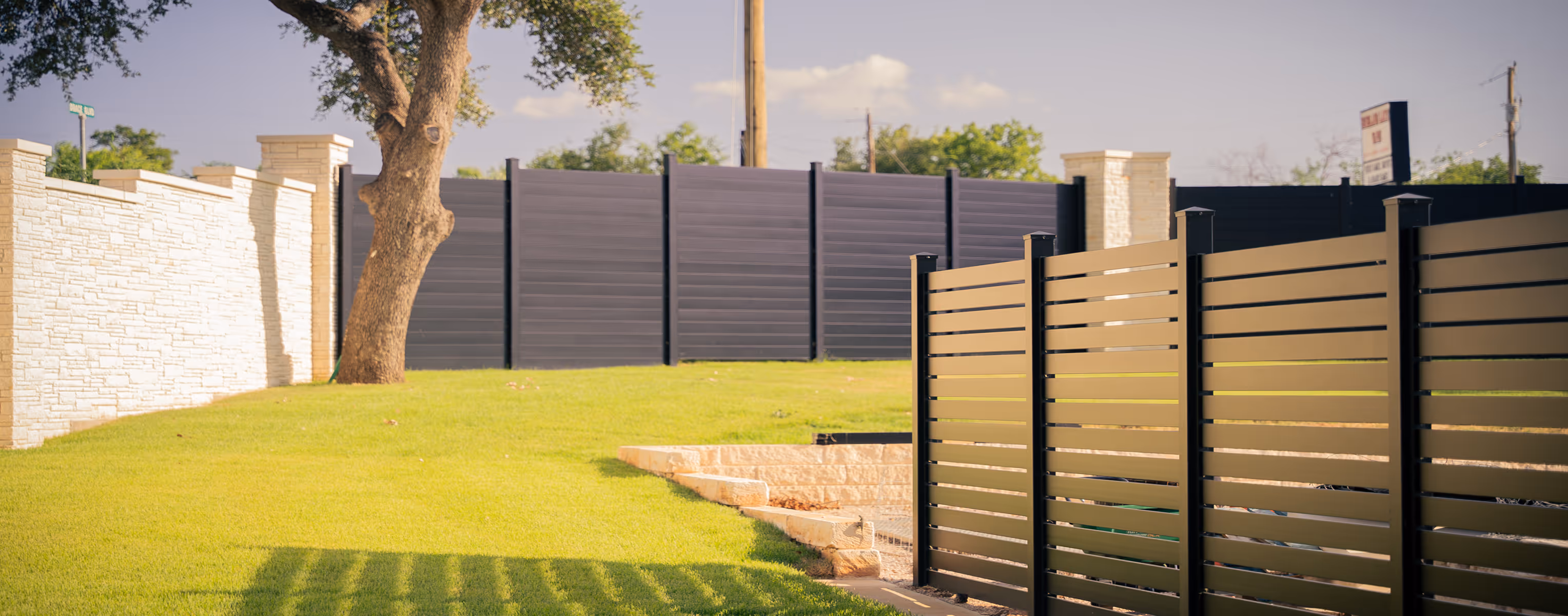 Green backyard with a large tree, white stone wall on the left, and modern horizontal slat fences in black and beige.