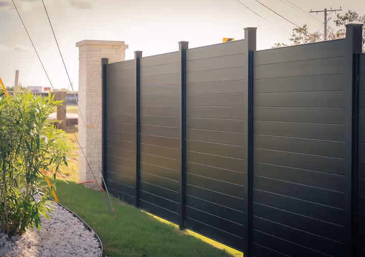 Modern black horizontal panel fence with a stone pillar on a lawn bordered by white rocks and green plants.