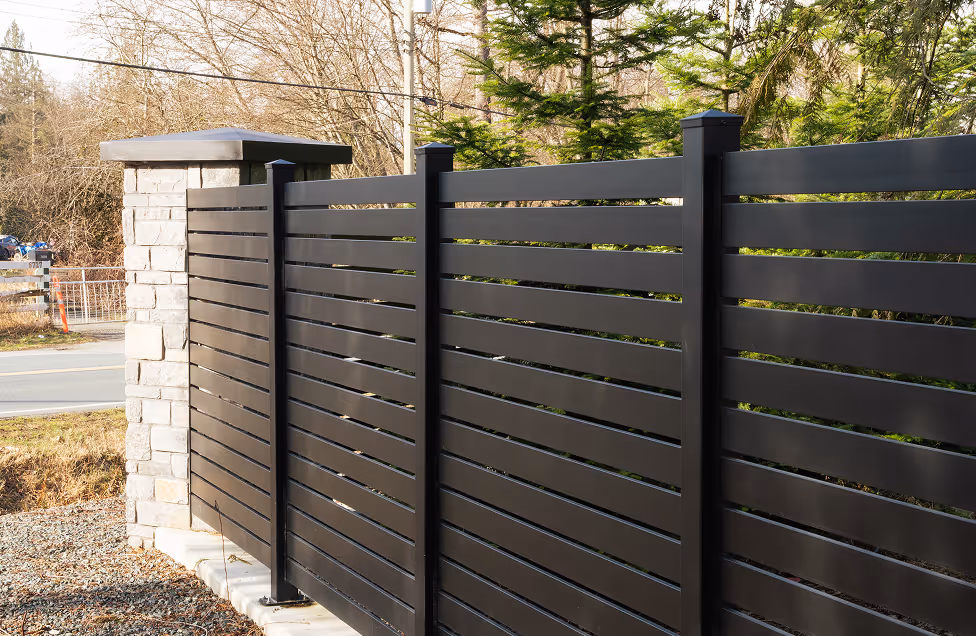 Modern black horizontal slat metal fence with a stone pillar at the corner surrounded by greenery.
