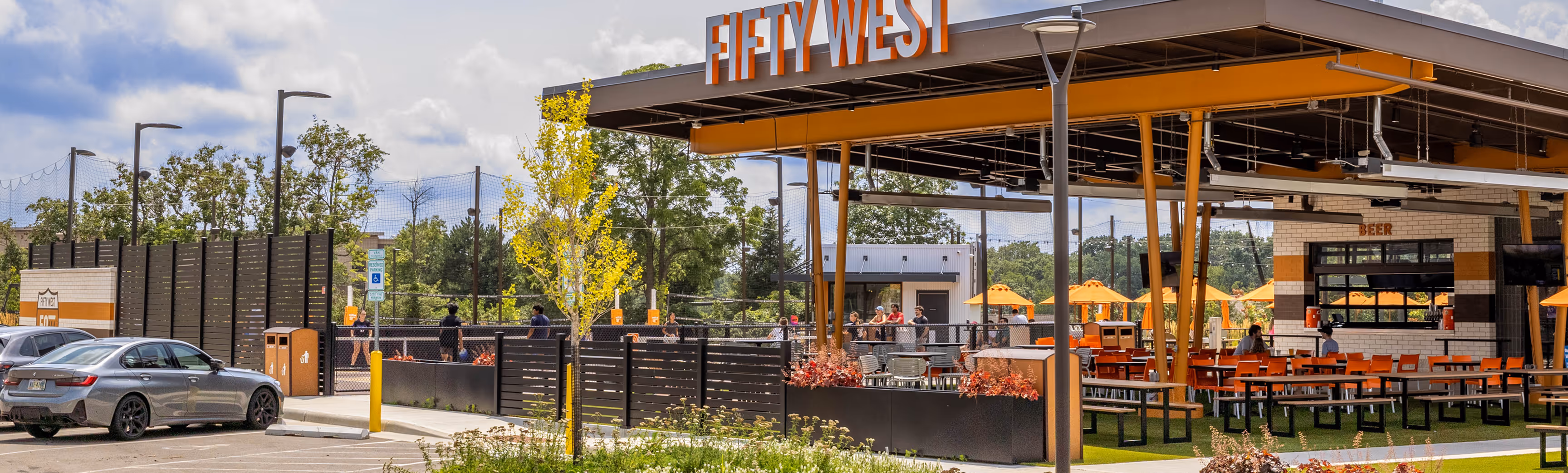 Outdoor seating area of Fifty West brewery with orange chairs, tables, and umbrellas next to a parking lot and people walking near a fence.