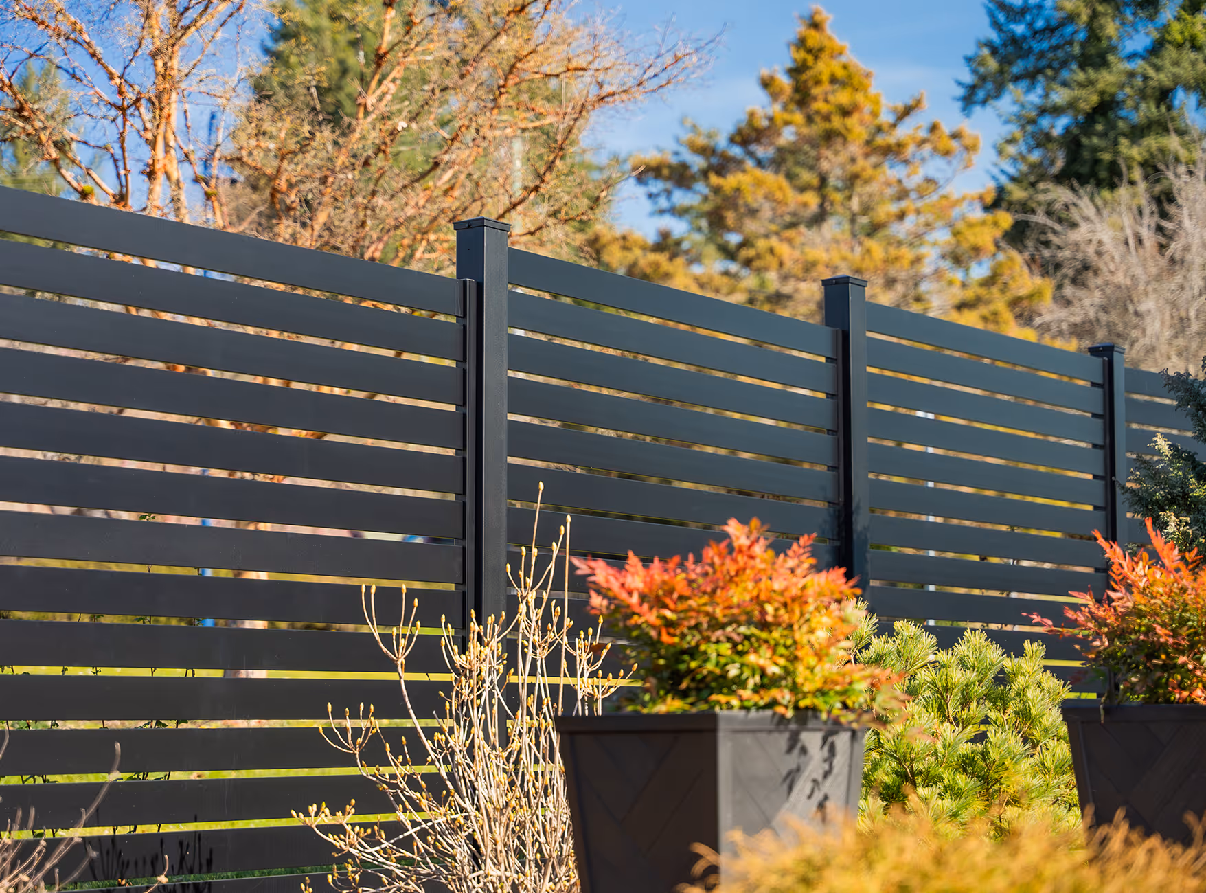 Modern black horizontal slat privacy fence with various green and orange shrubs in front under a blue sky.