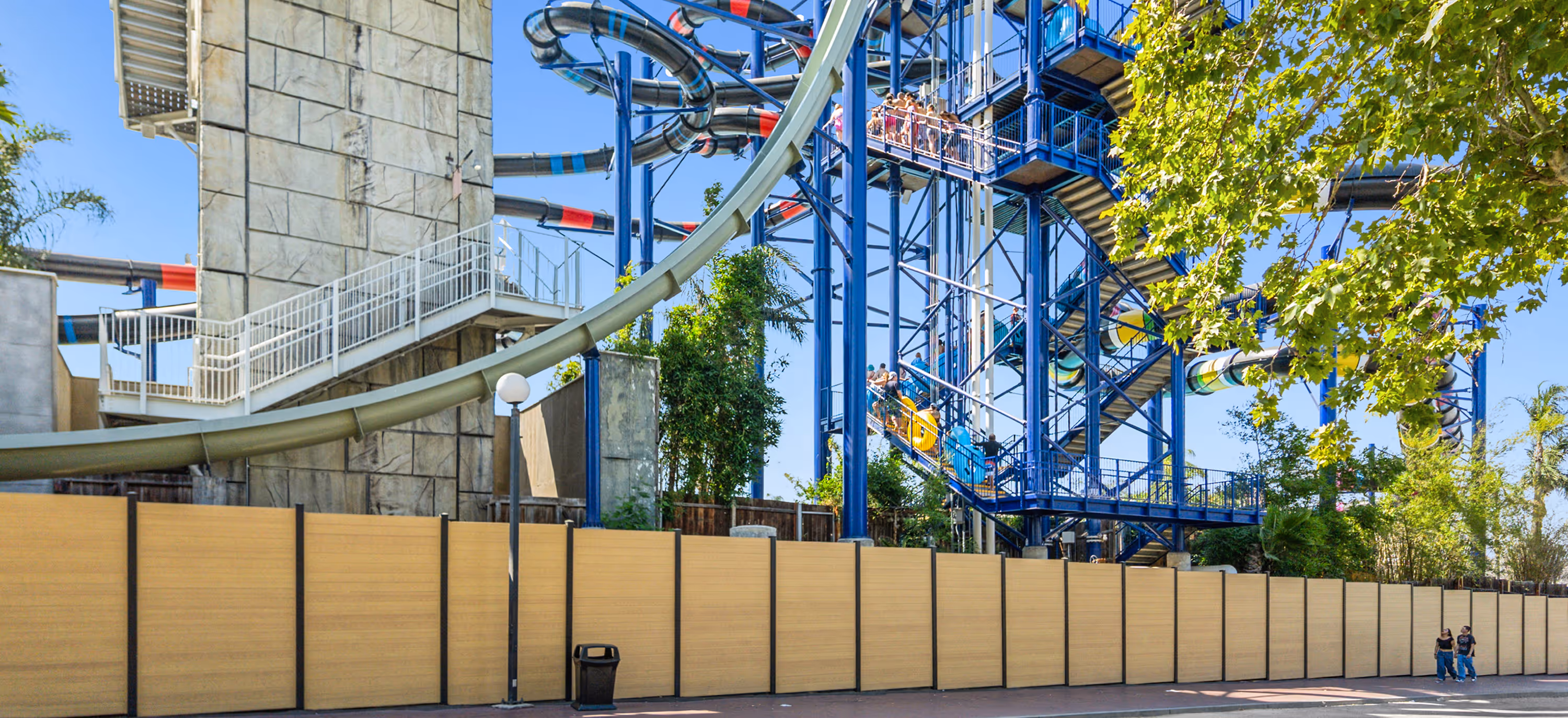 People queueing on blue metal stairs leading to a colorful enclosed water slide at a water park on a sunny day.