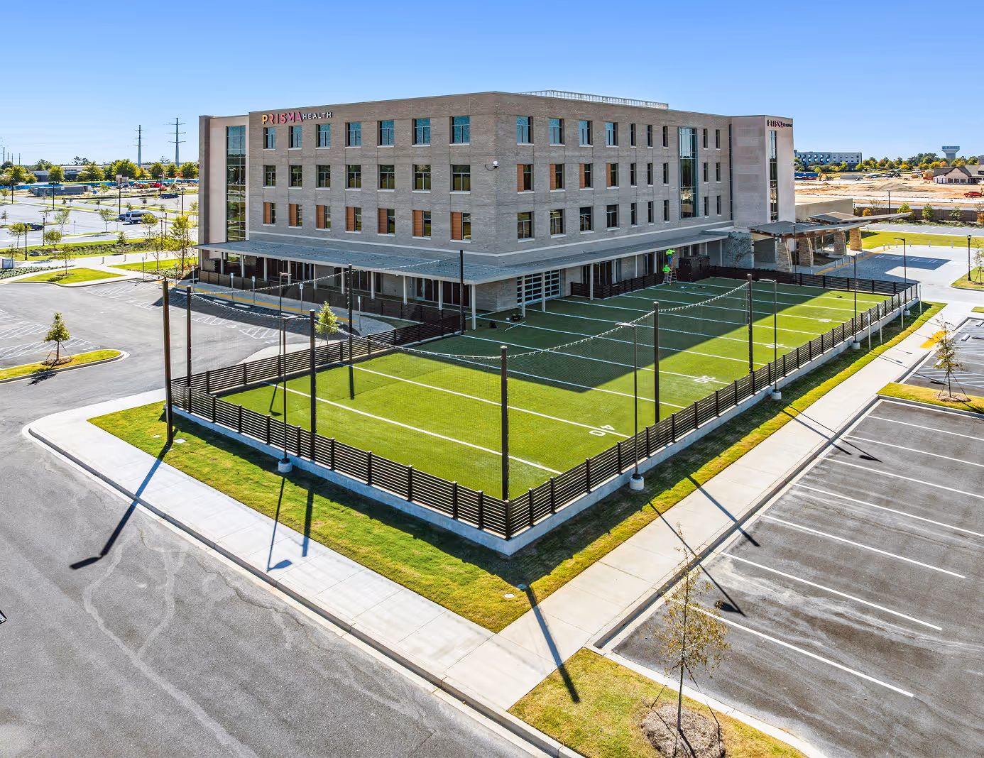 Four-story Prisma Health building next to a fenced artificial turf sports field and empty parking lots under a clear blue sky.